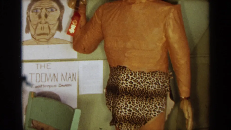 1963 Texas 8mm frame showing a homemade caveman figure with club, leopard-print costume, green chair, and hand-drawn evolution posters.