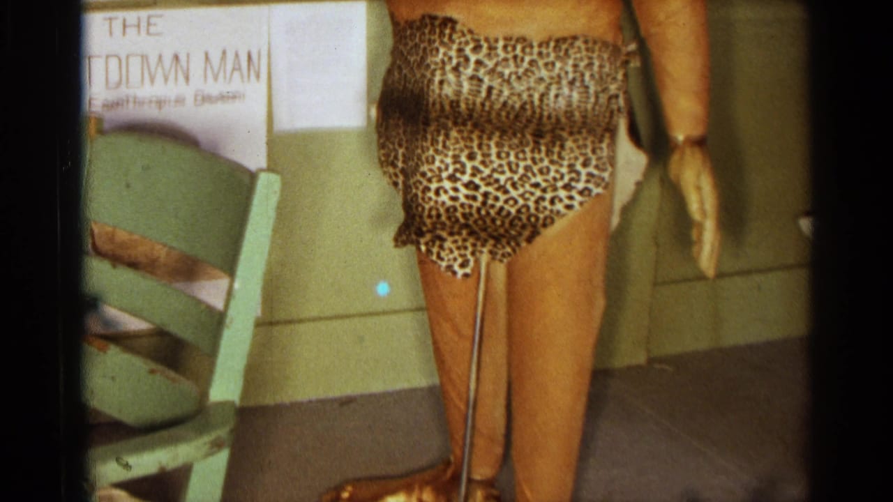 Lower half of caveman statue with leopard-print costume and gold-colored shoes near a chair.