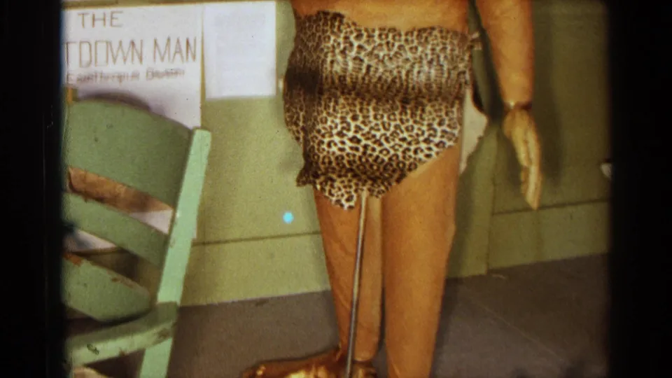 Close view of the caveman figure's leopard-print loincloth, bare legs, support rod, gold-colored feet, and green chair in the display room.