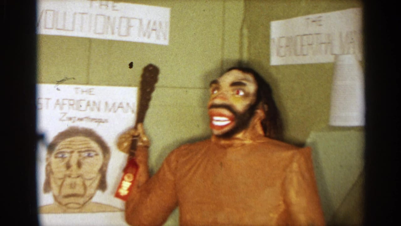 Texas 1963 home-movie frame of a caveman statue with painted face, raised club, and surrounding hand-lettered evolution display materials.