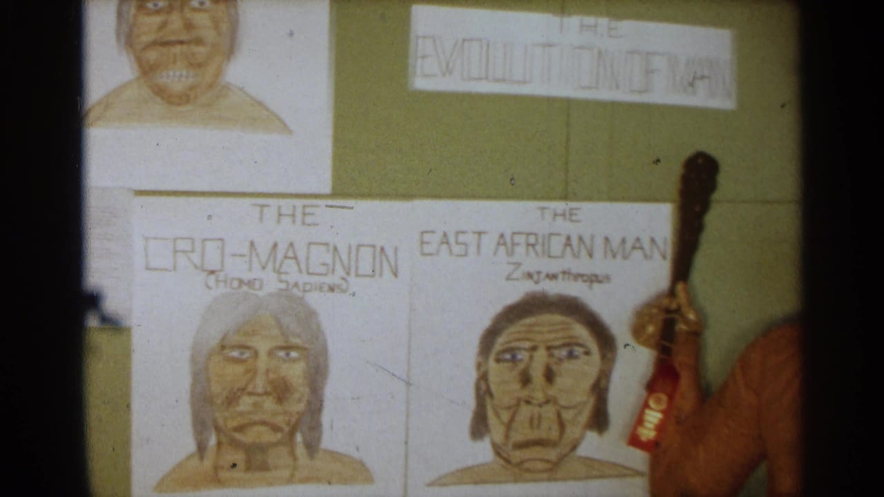 Hand-drawn evolution posters labeled Cro-Magnon and East African Man appear on a green wall beside the edge of the caveman statue and club.