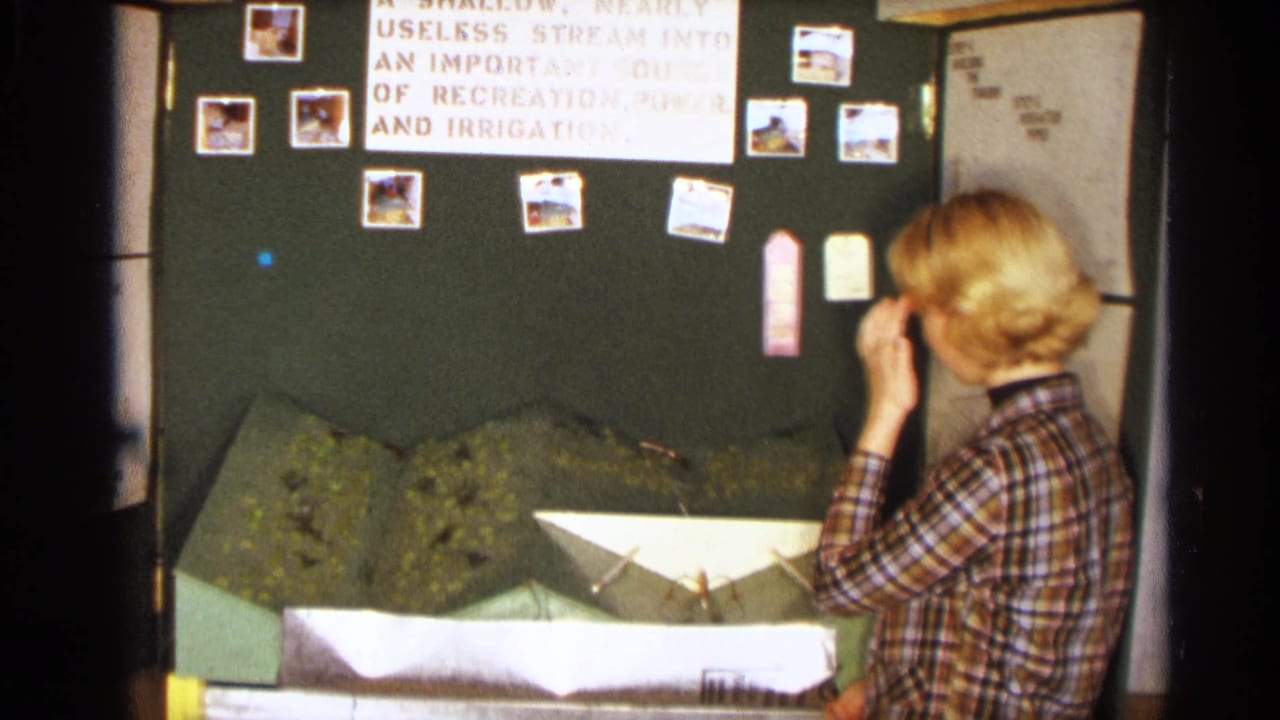 The girl stands near the model dam display in the Texas classroom, looking toward labels and photographs on the board.