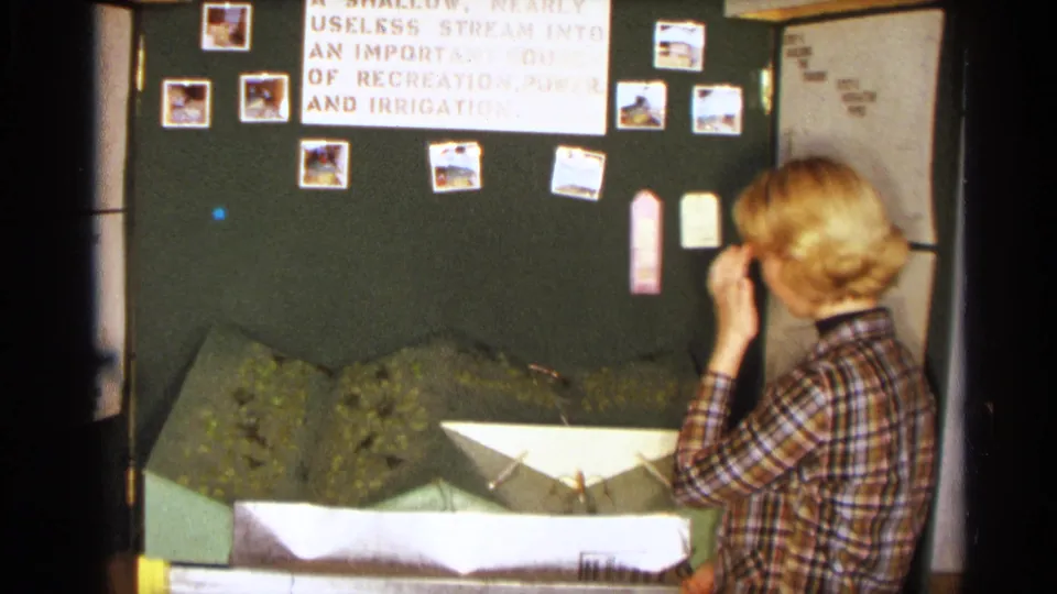 The girl stands near the model dam display in the Texas classroom, looking toward labels and photographs on the board.
