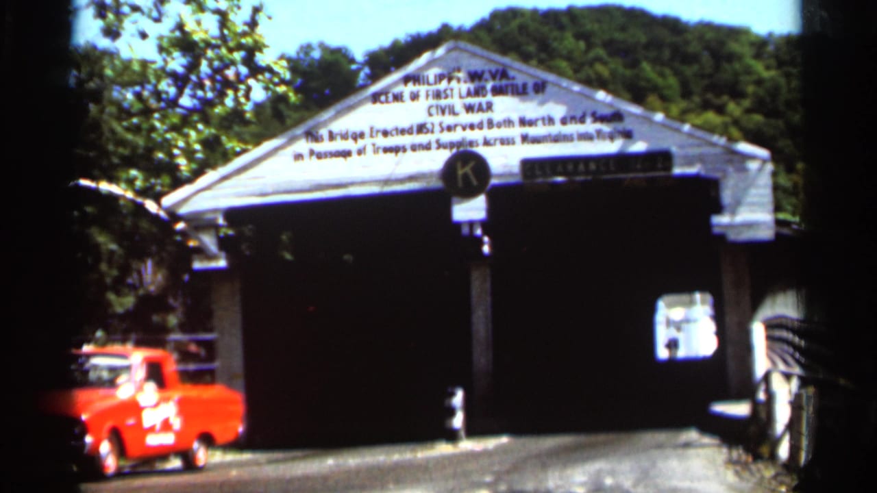 Covered bridge entrance with white gable sign, trees, and a red vehicle at left.