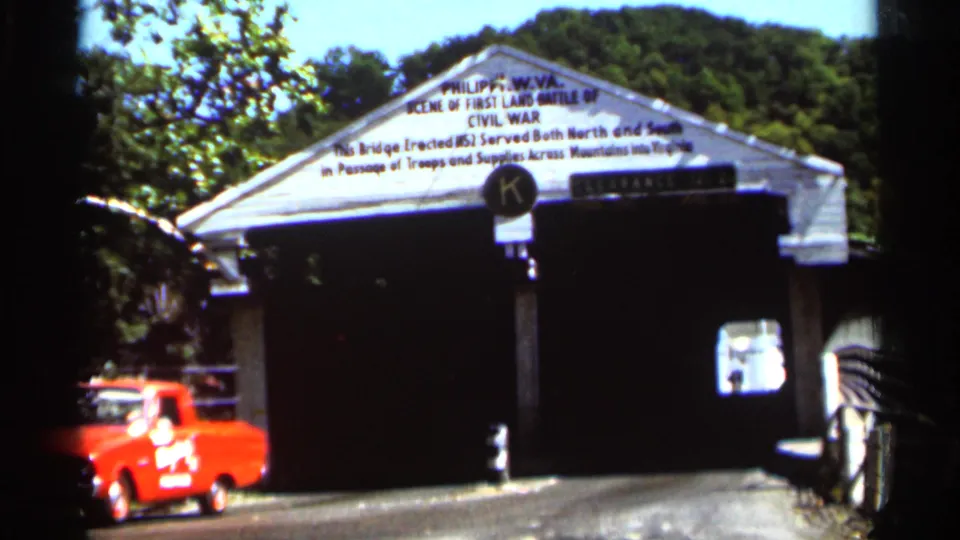 1964 8mm frame showing a covered bridge entrance near Indianapolis, with a white gable sign, wooded background, and red vehicle.