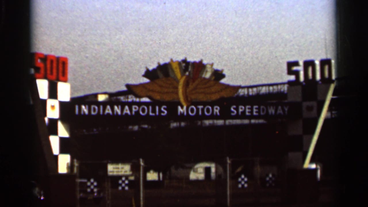 1964 8mm view of the Indianapolis Motor Speedway gate, centered on the sign, winged emblem, checkered decoration, and entrance fence.