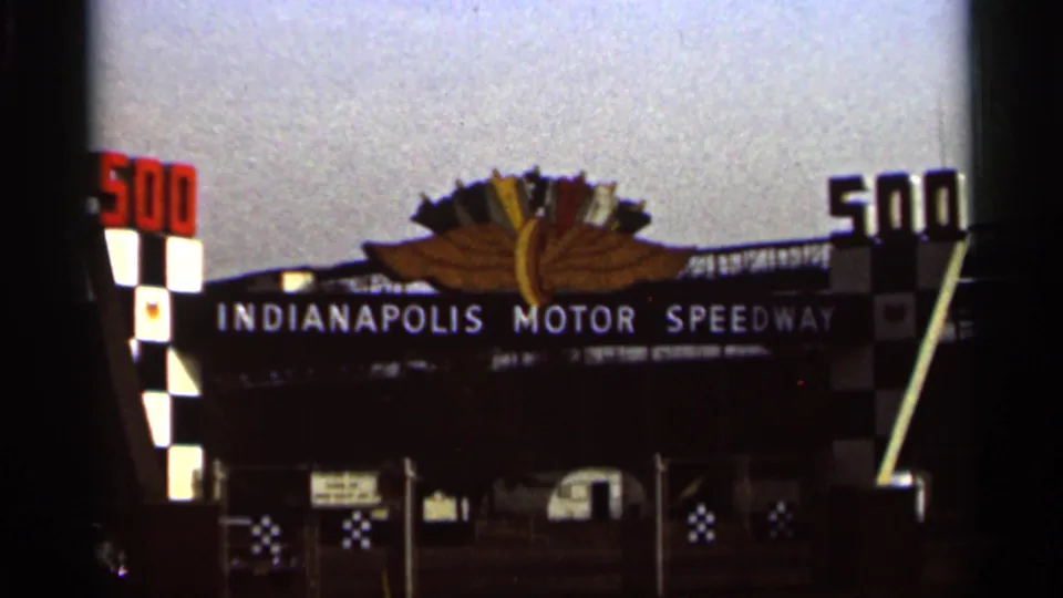 1964 8mm view of the Indianapolis Motor Speedway gate, centered on the sign, winged emblem, checkered decoration, and entrance fence.