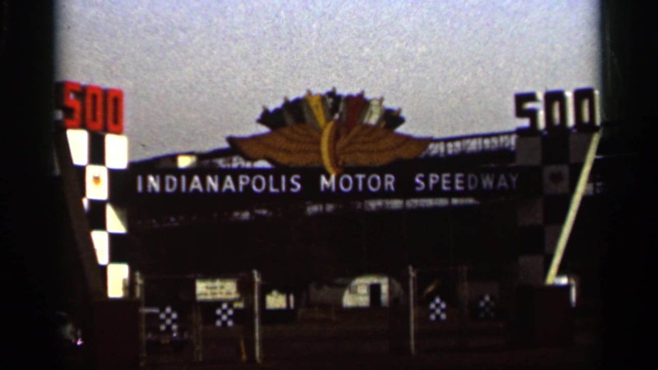 1964 8mm frame showing the Indianapolis Motor Speedway entrance from a similar angle, with 500 signs and checkered gate features.