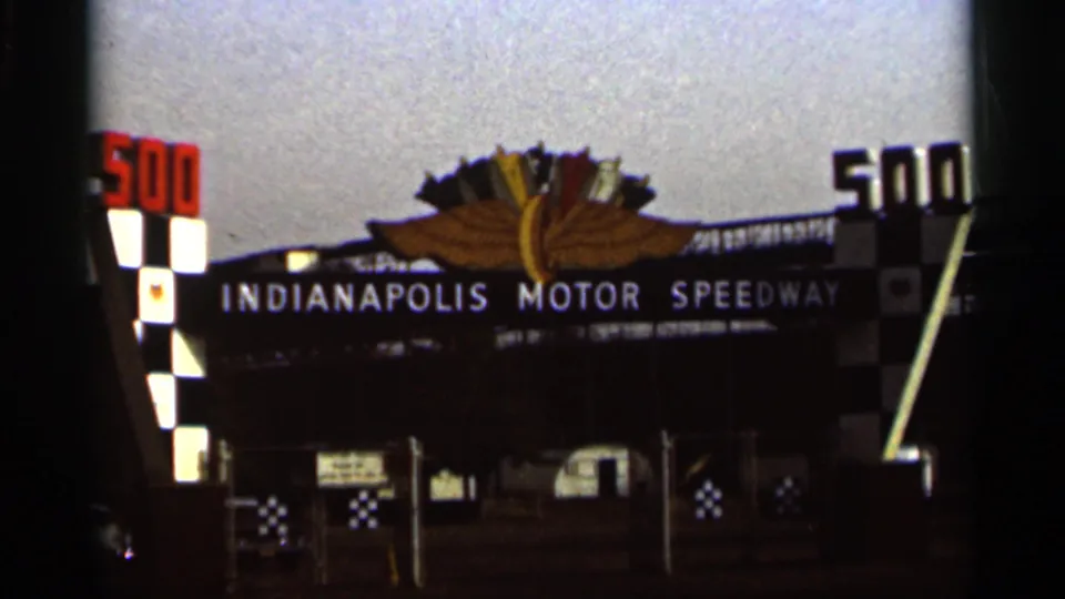 1964 8mm frame showing the Indianapolis Motor Speedway entrance from a similar angle, with 500 signs and checkered gate features.