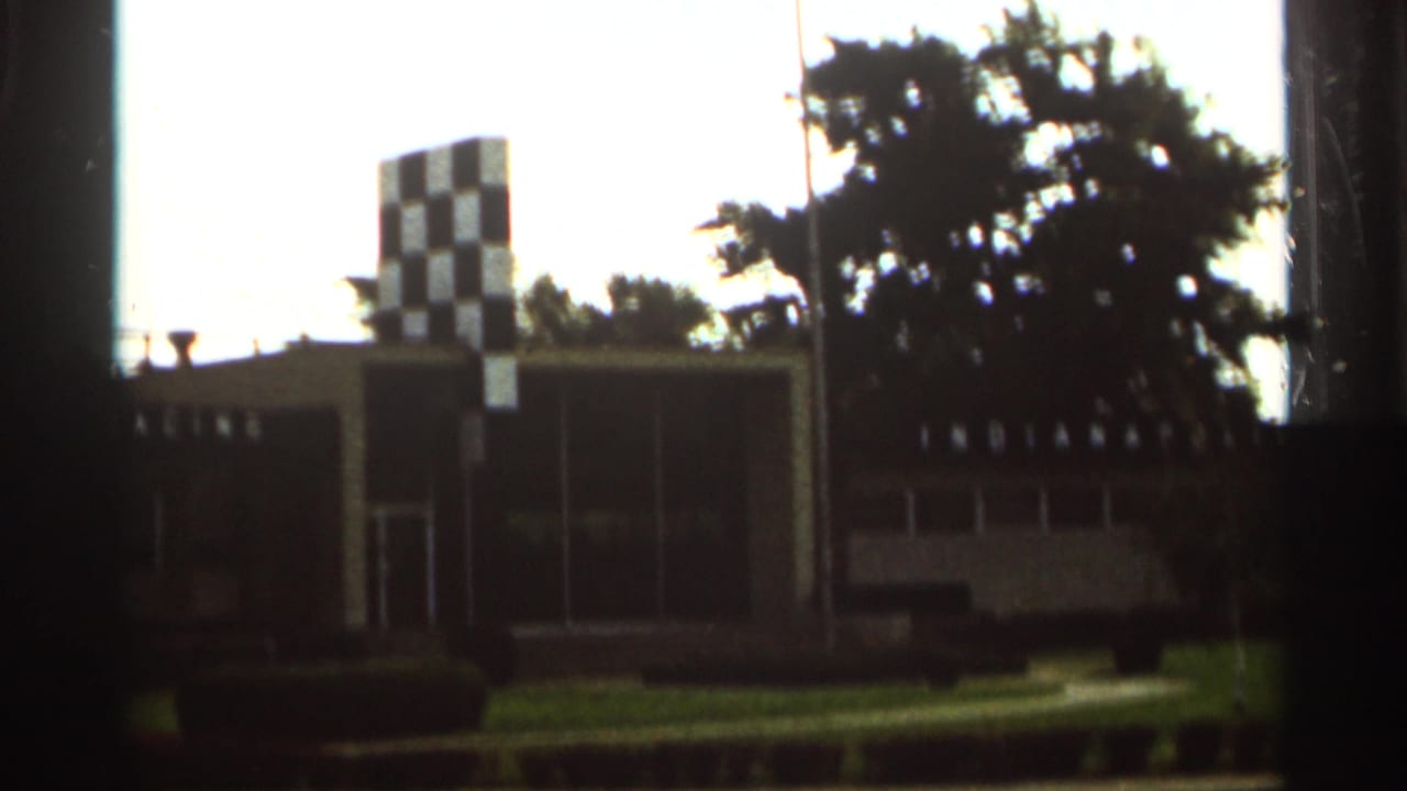 1964 Indianapolis Motor Speedway view with the checkered tower, low buildings, grass, and trees.