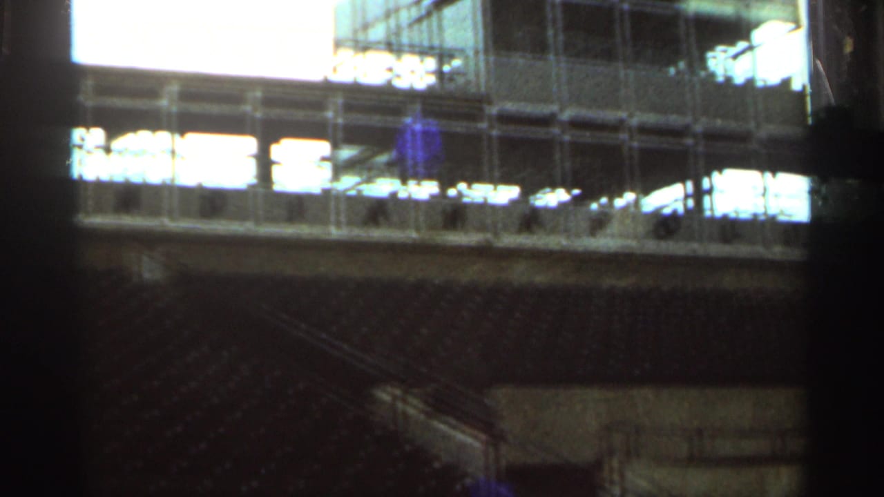 1964 Indianapolis Motor Speedway frame showing empty seating, railings, and a glass-fronted trackside structure.