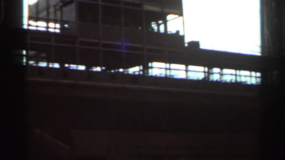 1964 8mm frame from Indianapolis showing a shadowed grandstand or trackside structure with bright daylight beyond.