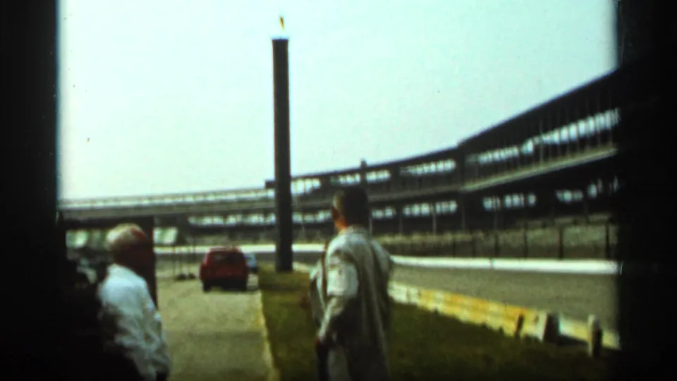 Two adult men stand along an Indianapolis race-track lane in 1964, with a red car, barriers, and grandstand structures visible.