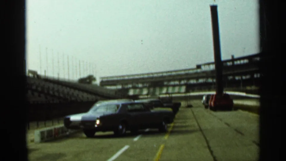 A dark classic car sits on or beside the Indianapolis track lane, with grandstand seating and a tall pole in the background.