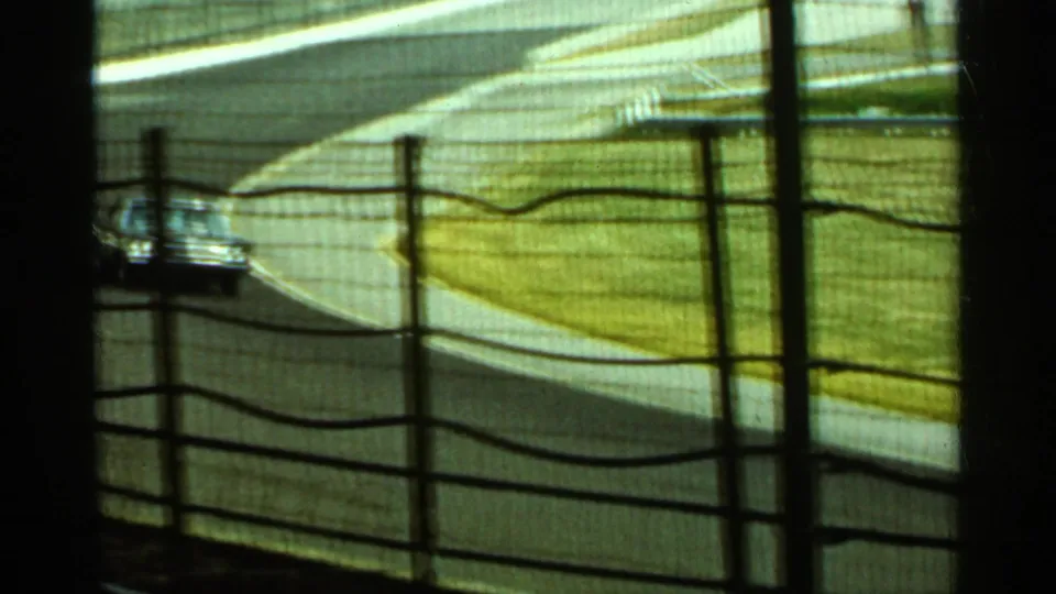 Yellow-green 8mm frame of a roadway and car beyond a wire fence, likely in the Indianapolis Gasoline Alley setting.