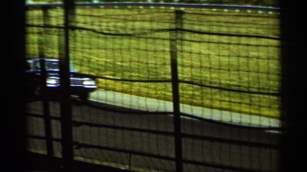 A dark race car moves along an Indianapolis racetrack curve in 1964, seen through protective fencing and heavy 8mm film grain.