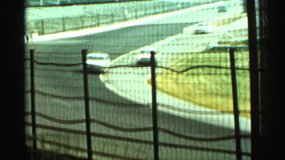 A light-colored race car rounds a sunlit Indianapolis track bend in 1964, partially obscured by protective fencing.
