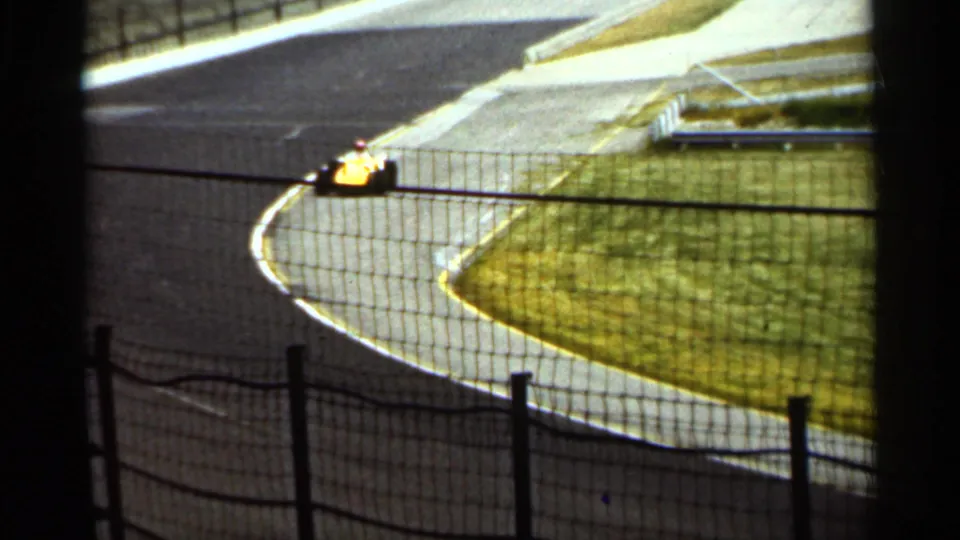 A yellow race car appears low in frame while taking a 1964 Indianapolis racetrack curve, with fencing and infield grass visible.