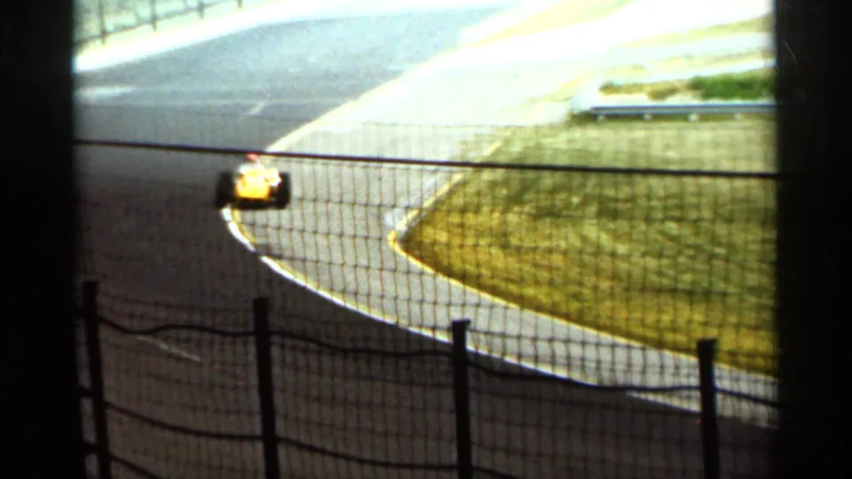 A yellow race car continues around the Indianapolis racetrack curve in 1964, softened by bright exposure and 8mm grain.