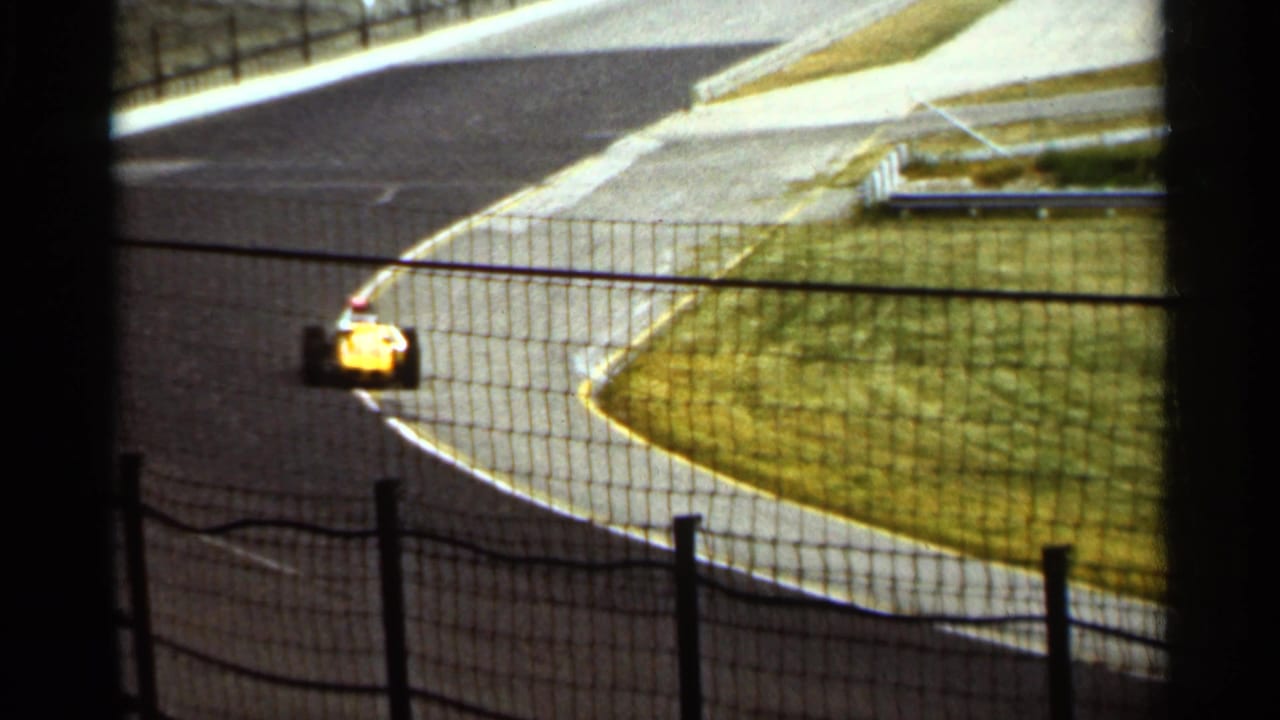 A yellow race car rounds a paved curve at Indianapolis Motor Speedway in 1964, seen from behind dark safety netting and posts.