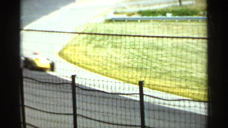 A yellow open-wheel race car appears blurred by motion on a sunlit Indianapolis track curve, with grass and safety fencing in view.