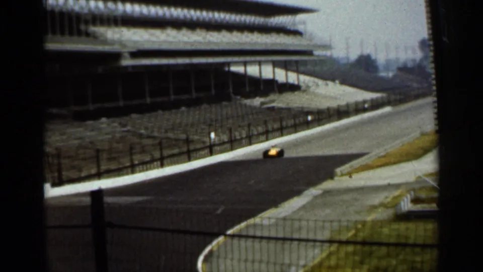 A distant yellow race car travels along the Indianapolis speedway surface, with grandstand roofing, fencing, and hazy background visible.