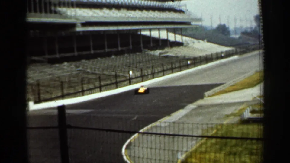 A small yellow race car continues along a curved Indianapolis track section, framed by safety netting, fencing, grass, and grandstand edges.