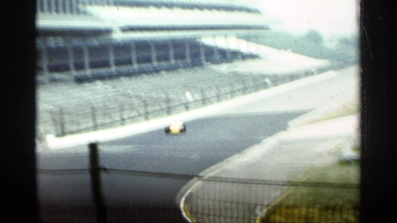 Yellow race car entering a racetrack curve behind foreground fencing.