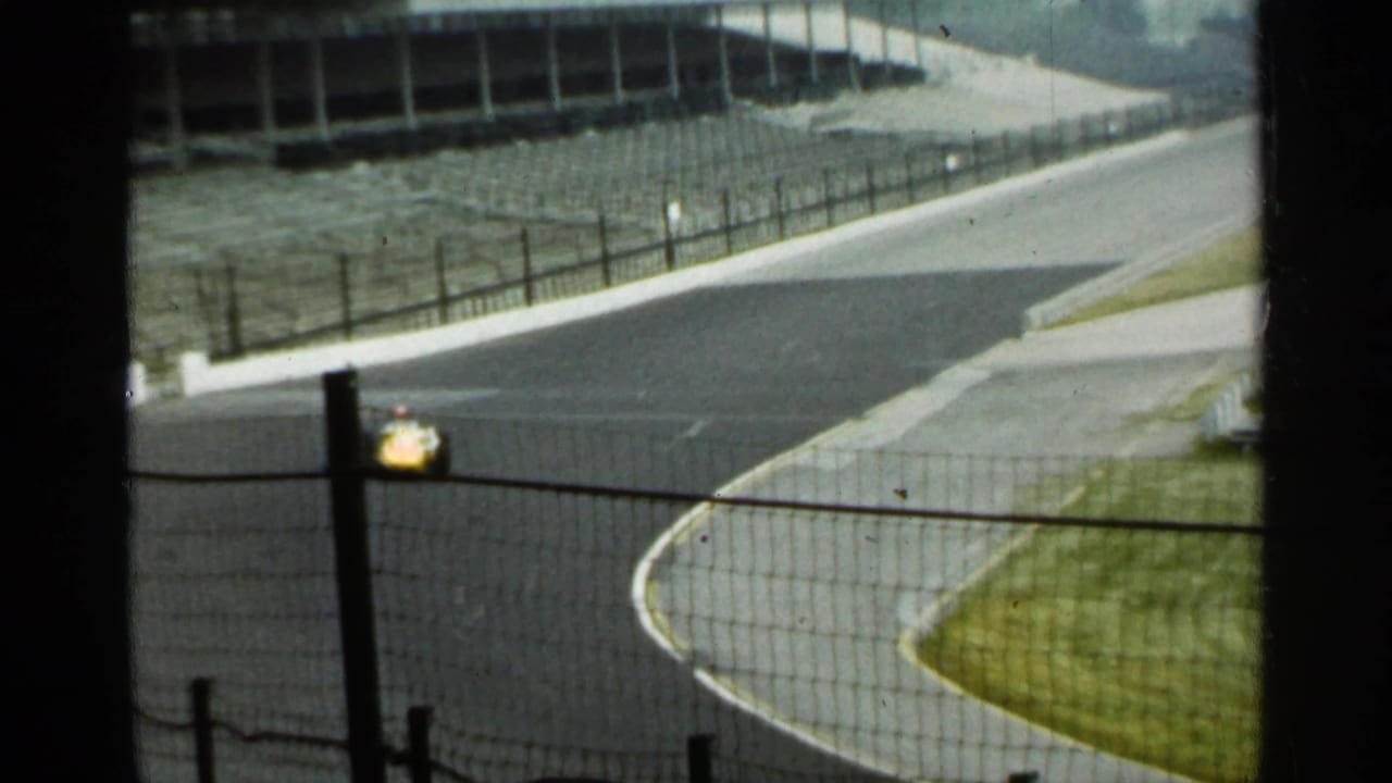 Yellow race car on a paved track curve with empty grandstands in the background.