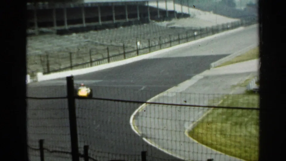 A yellow race car travels along the Indianapolis racetrack curve, with a white wall, fencing, and empty grandstands visible.