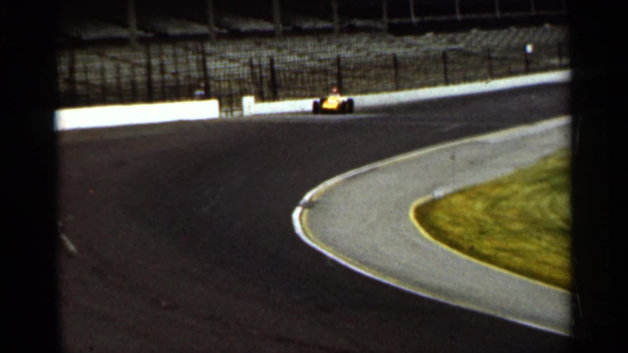 The yellow car rounds the Indianapolis track bend, framed by dark foreground fencing and the bright inner curb.