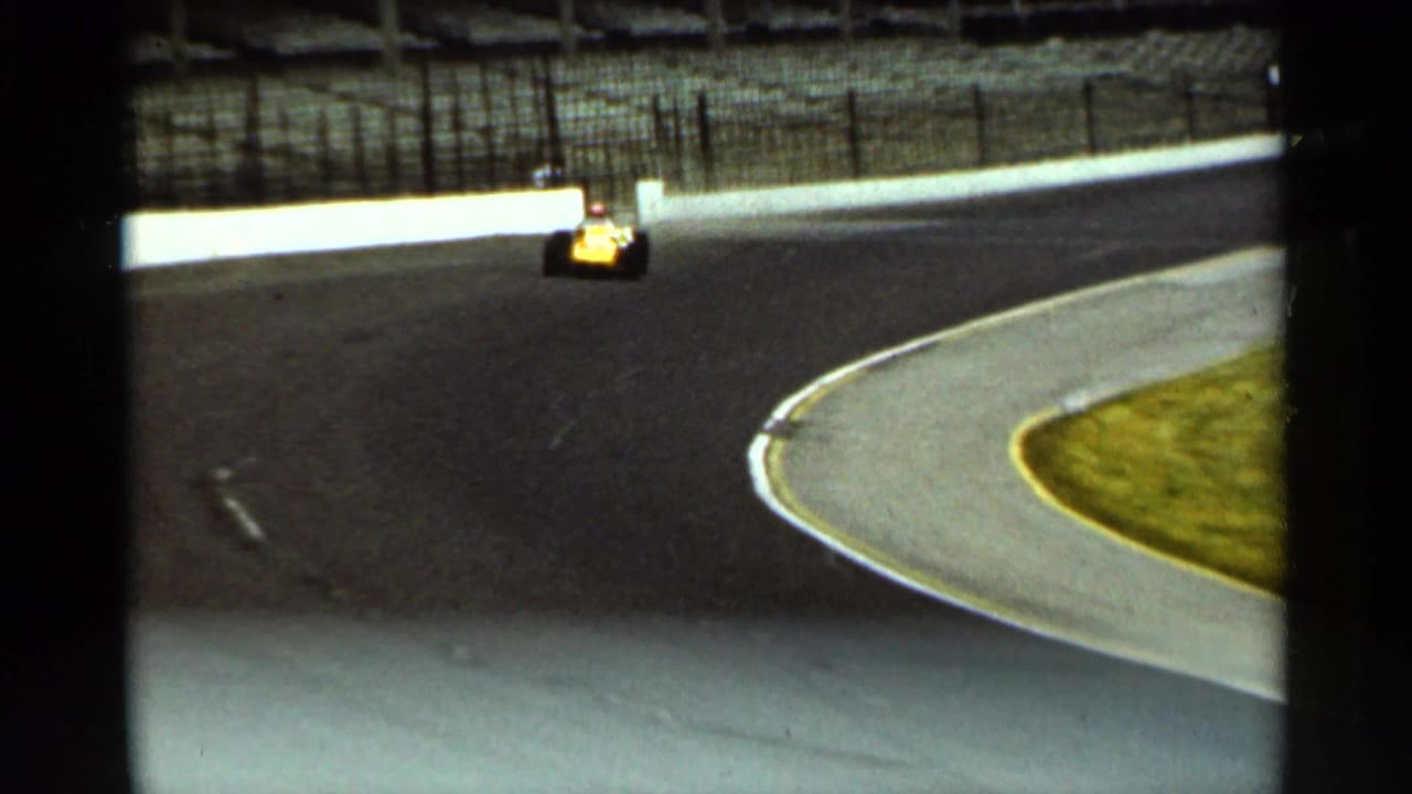 The yellow race car appears closer on the Indianapolis racetrack curve, with the paved turn, grass, and track wall visible.