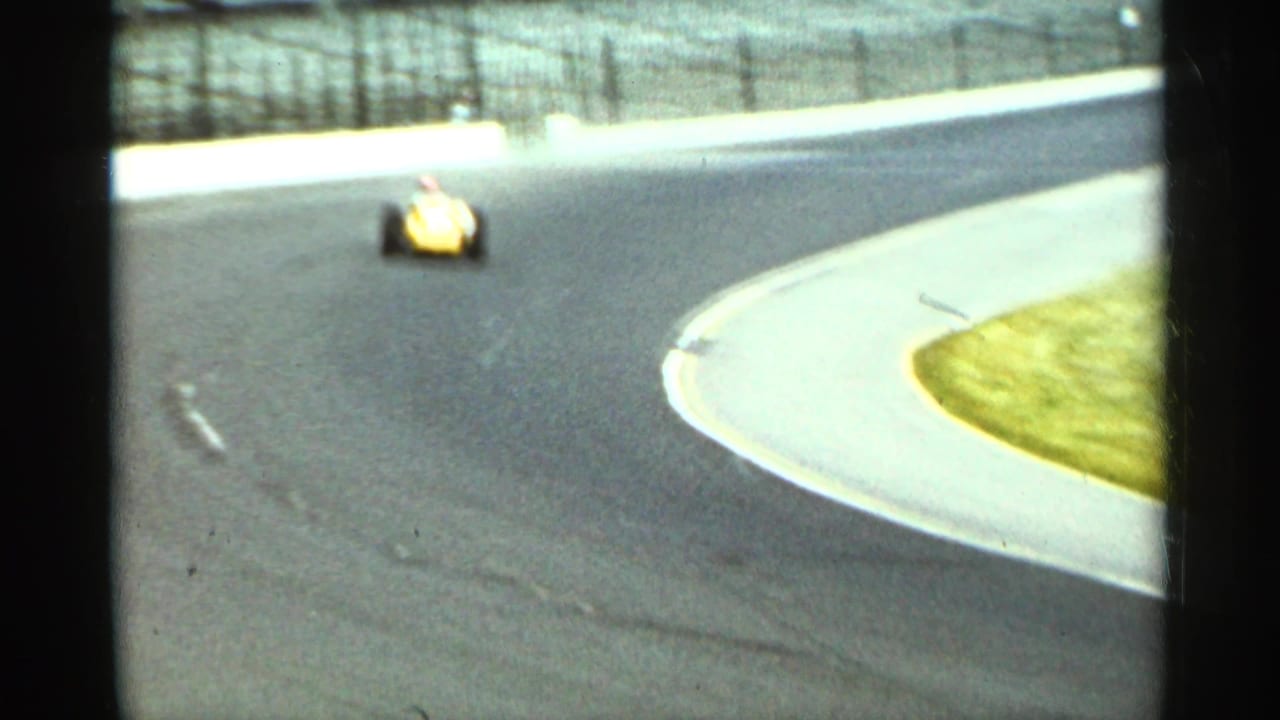 Yellow open-wheel race car approaches a paved track turn beside grass and fencing.