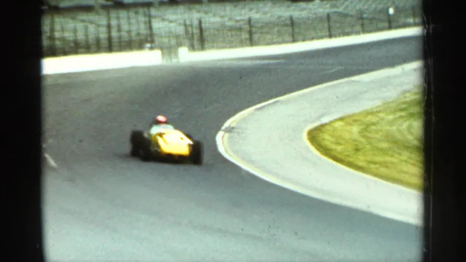 1964 Indianapolis home-movie frame with a yellow open-wheel car rounding a smooth track curve near pale curbing and grass.