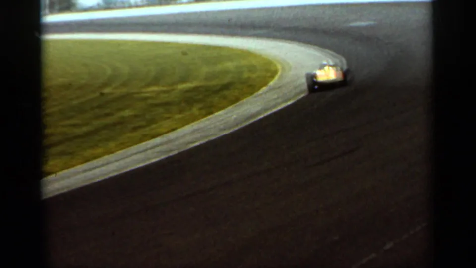 1964 Indianapolis 8mm frame showing the yellow race car farther through the bend, with grass infield and dark track surface prominent.