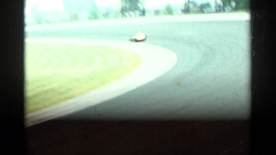 1964 Indianapolis 8mm frame showing the yellow open-wheel car small in the distance as it exits a bright track curve beside grass.