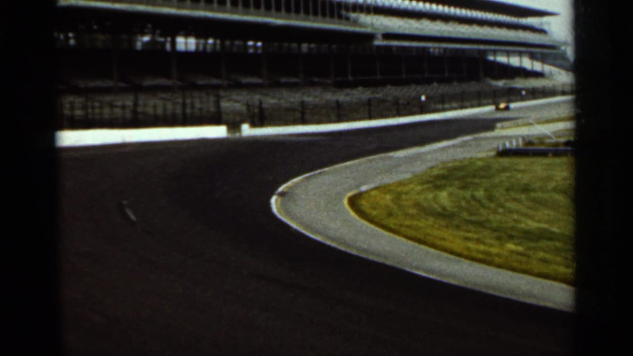 1964 Indianapolis speedway footage showing a sweeping track turn, grass infield, empty grandstands, and a distant race car.