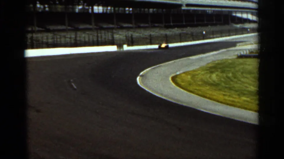 The race car continues around the sweeping Indianapolis turn, framed by the track apron, infield grass, and grandstand structure.