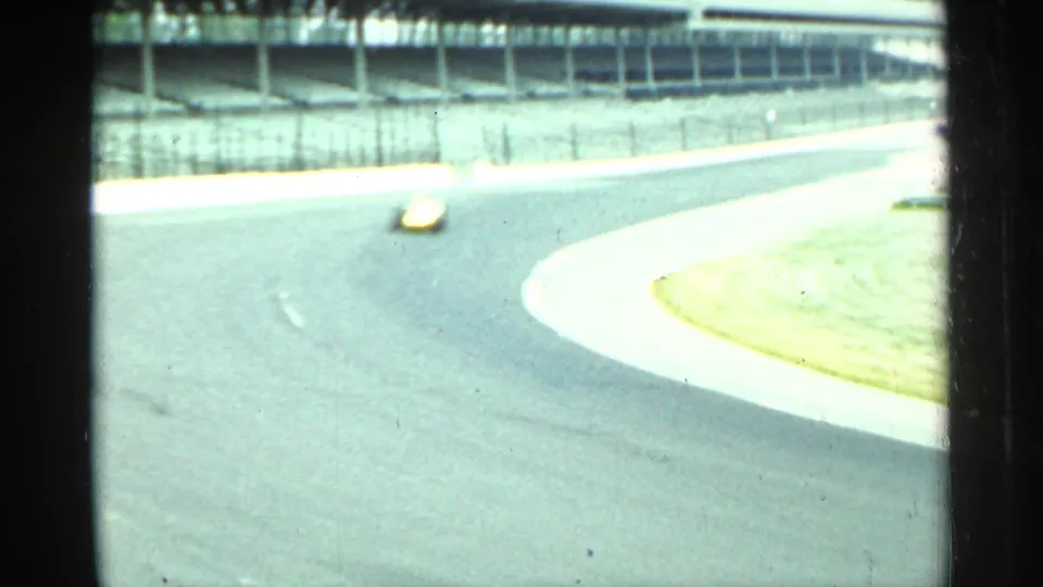 A brighter 8mm frame shows the yellow race car passing through the Indianapolis curve, with the track apron and grandstands washed by exposure.