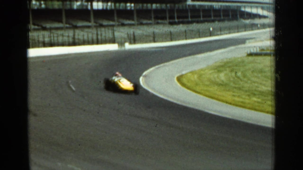 Yellow race car on the Indianapolis Motor Speedway track in 1964, blurred as it moves through a turn below the grandstands.