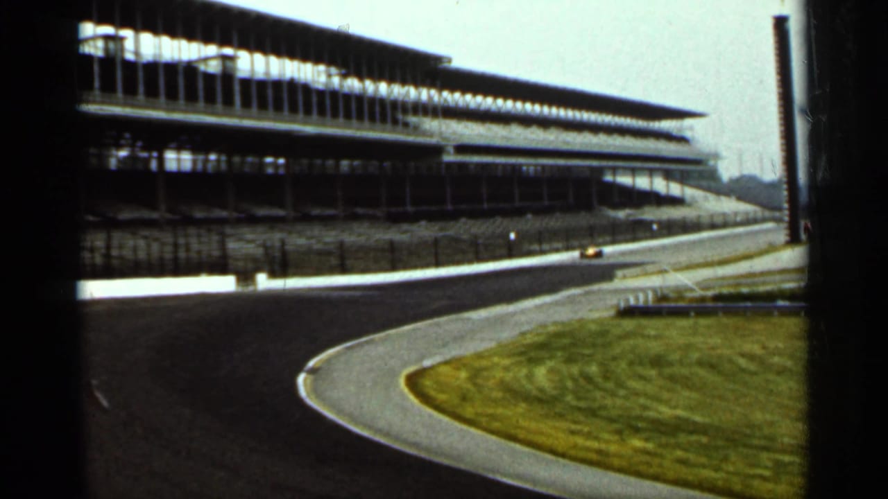 Wide view of the speedway turn with grandstands and a small yellow car in the distance.
