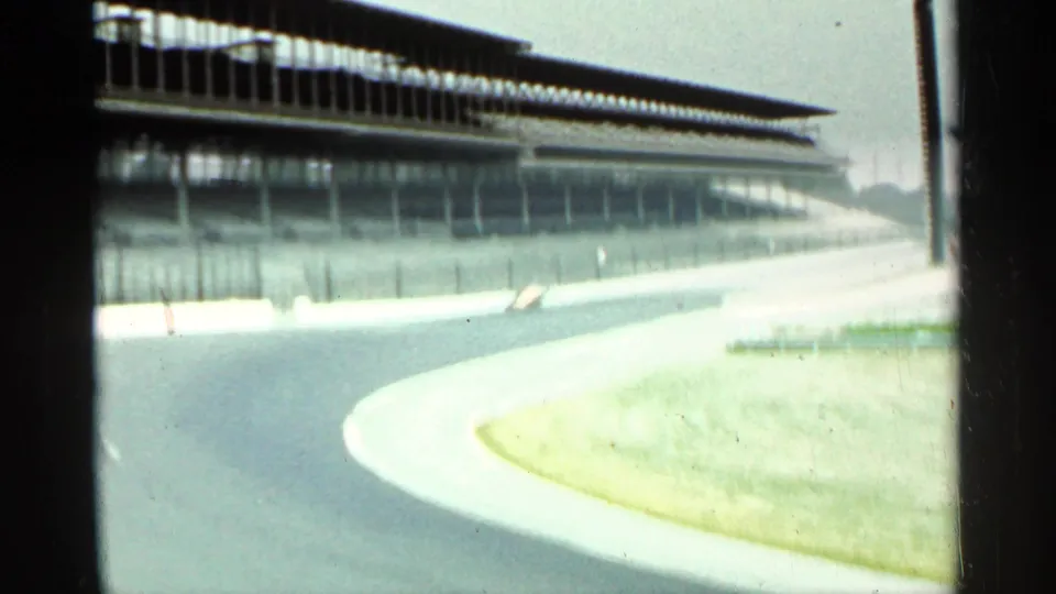 Bright, overexposed 8mm frame of the Indianapolis track curve in 1964, with grandstands, fencing, wall, and grass visible.