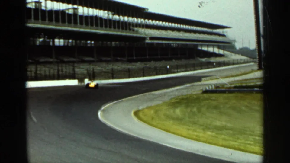 Yellow open-wheel car on the 1964 Indianapolis track, moving through the lower part of the turn below the grandstands.