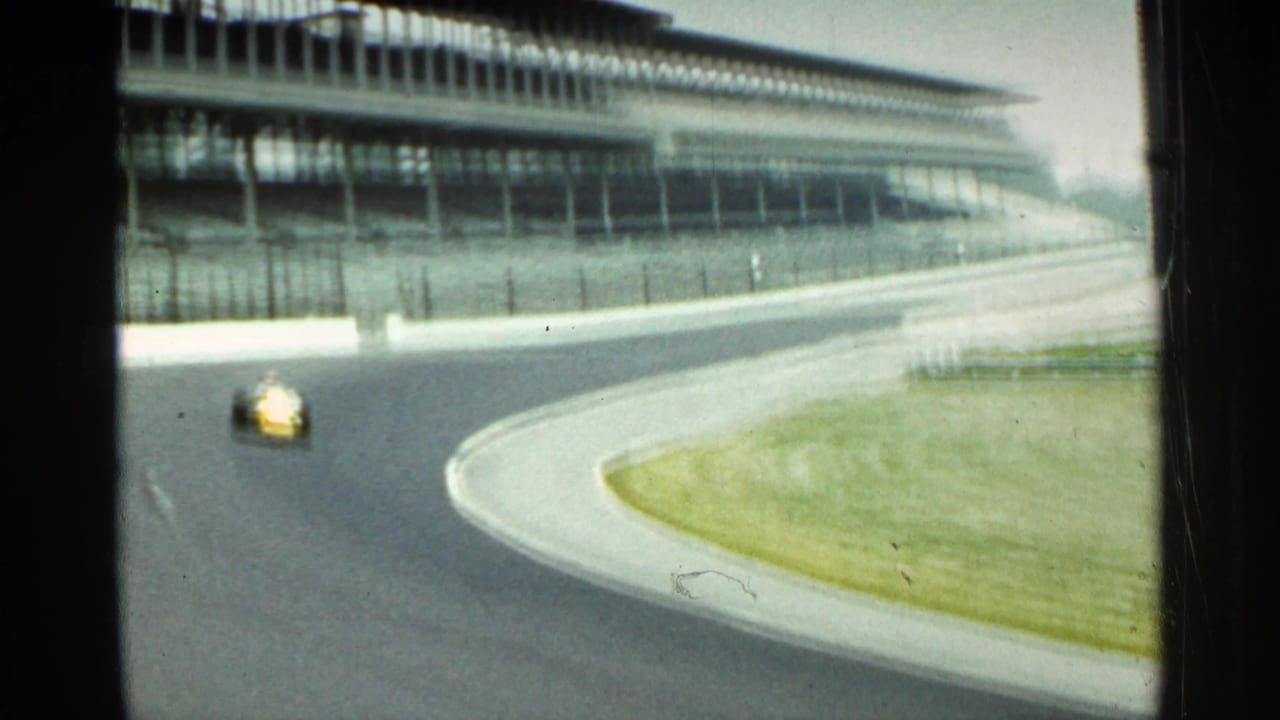 Yellow race car rounding a speedway turn near large grandstands.
