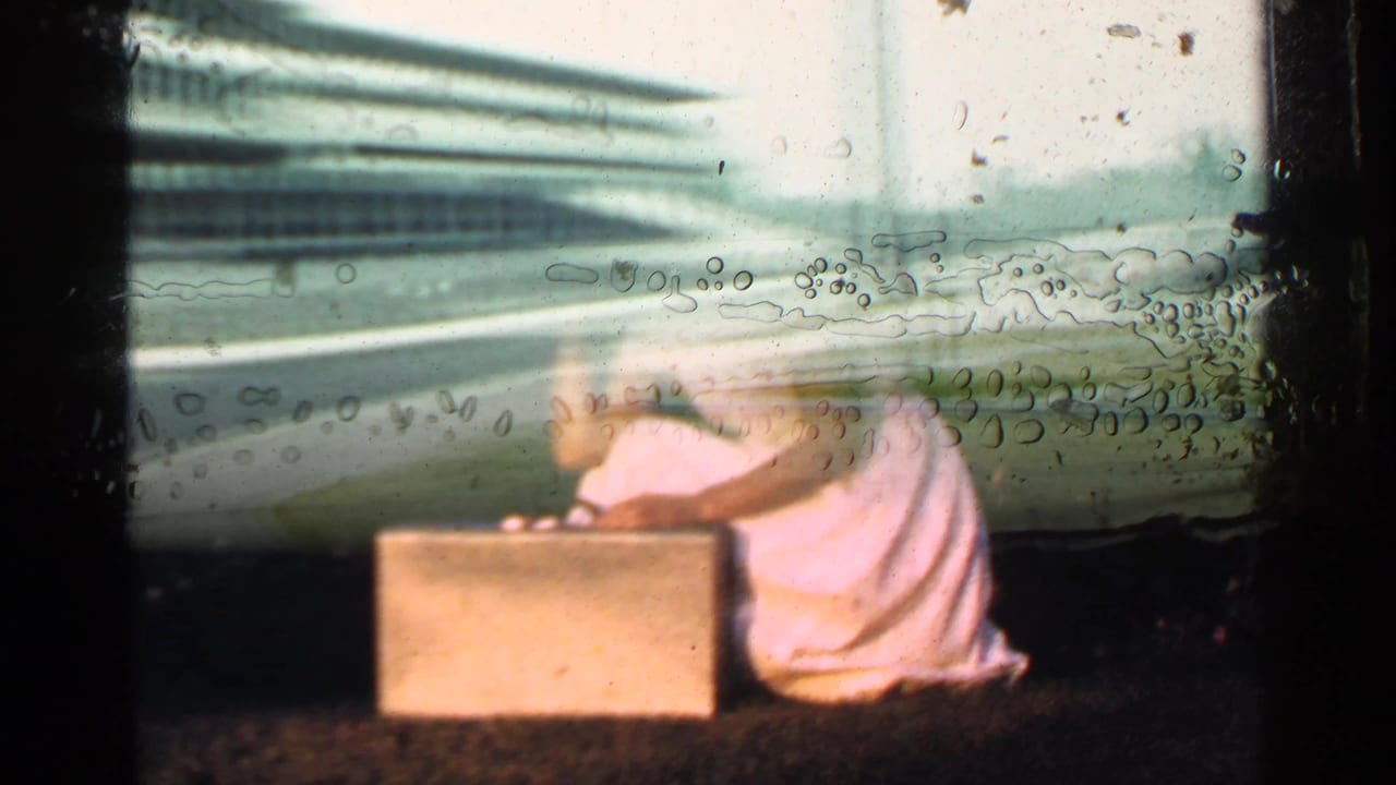 A woman in a white dress kneels on a lawn beside a rectangular briefcase, seen through faded 8mm home-movie color.