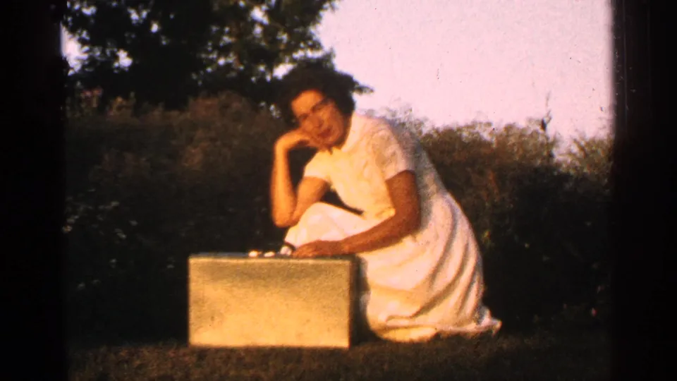 The woman in a white dress sits or kneels on grass, leaning on one hand while positioned behind a briefcase in an outdoor yard.