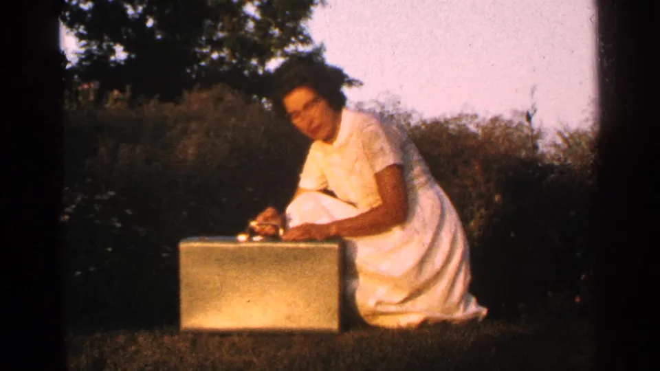 A woman in a white dress kneels outdoors and appears to handle items on a rectangular briefcase in a 1960s home-movie scene.
