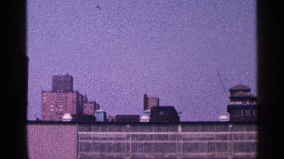 1964 New York City home-movie frame showing brick rooftops and apartment buildings through soft 8mm grain.