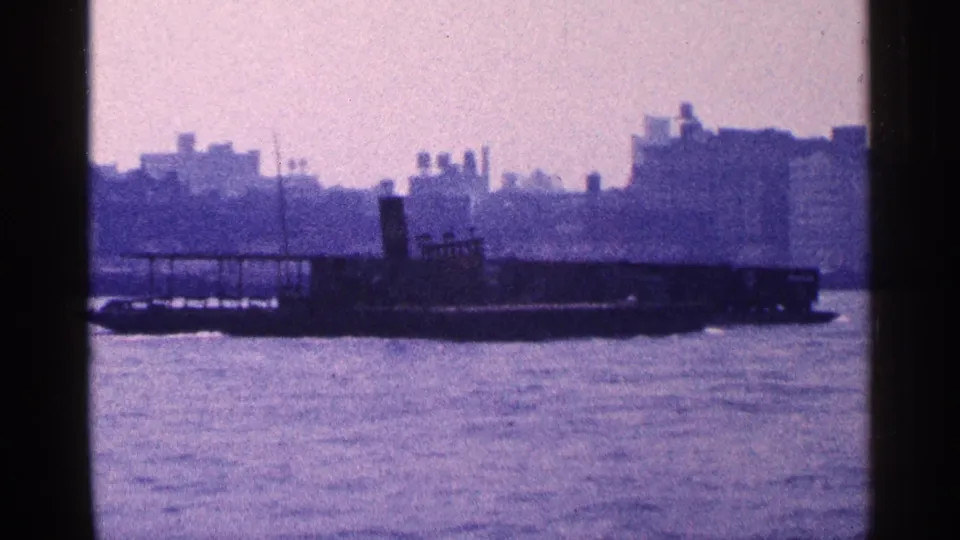 1964 New York City waterfront frame showing a dark barge crossing water before a hazy urban backdrop.