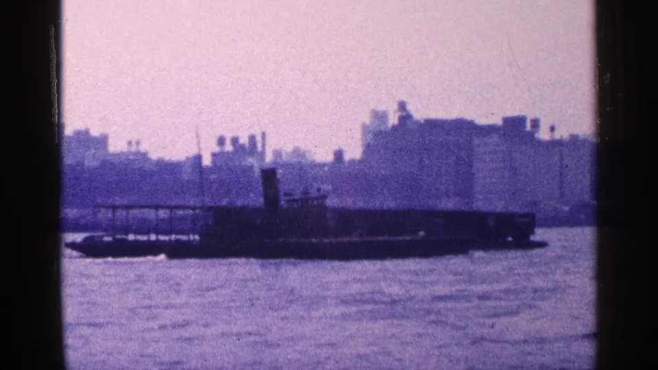 1964 New York City home-movie frame of a barge on the water against a soft, distant urban shoreline.